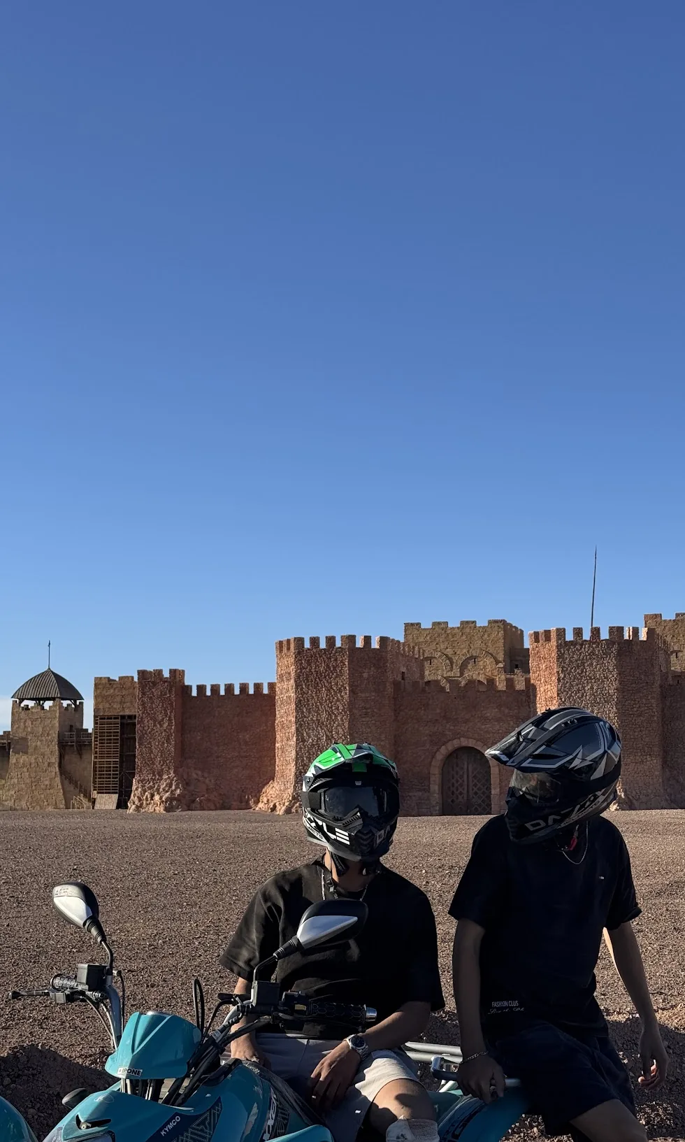 Two helmeted riders on quads in front of a long fortified wall